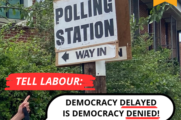 Caption in front of a polling station sign: Tell Labour Democracy Delayed is Democracy Denied.